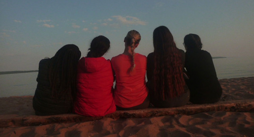 Five people sit close together with their backs facing the camera. They are looking out over a body of water. 